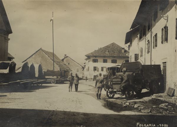 Street view of Folgaria in South Tyrol. Soldiers are walking down the street, and two other soldiers are near a truck on the right. Red Cross wagons are parked in a row along the side of the street on the left. The flagpole also has a Red Cross flag. 
