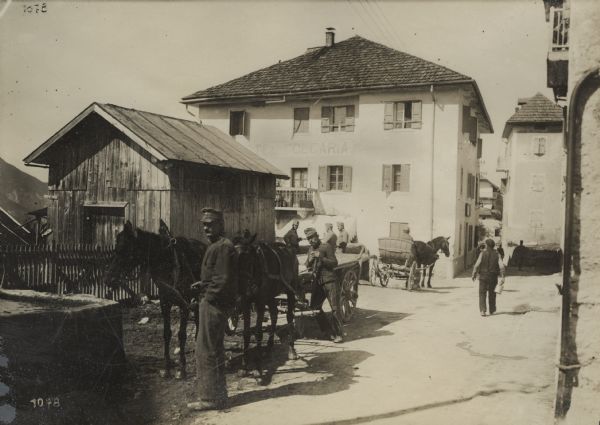 Soldiers relaxing in front of the Folgaria Hotel in Italy. The men are standing around horse-drawn wagons and carts, and other men are walking down a street or alley near the hotel. There is a mountain in the background on the left. 
