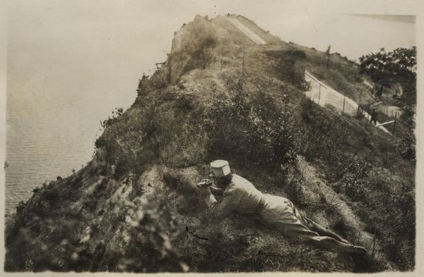 View looking down at an Austrian fortress officer lying in the grass at the crest of a ridge line, using it as an observation post while looking for the enemy above a lake on the southwestern battlefront in Tyrol.