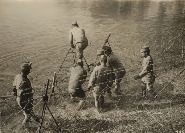 Austrian soldiers are setting up a barbed wire defense on a shoreline in the South Tyrol. The men are barefoot and standing in the water, while one of the men is standing on a ladder.