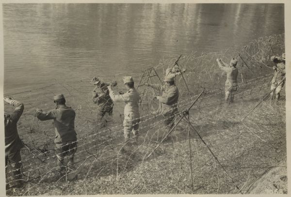 Austrian soldiers are setting up a barbed wire defense on the shoreline. The men are barefoot and standing in the water with their pants rolled up.