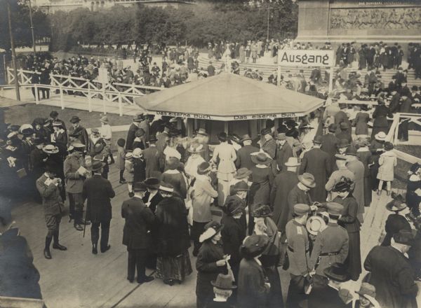 The life and hustle and bustle at the "Iron Hindenburg" monument. The military band playing in front of the statue. People are gathered around a booth to make donations for the privilege of driving iron nails into the wooden statue. 