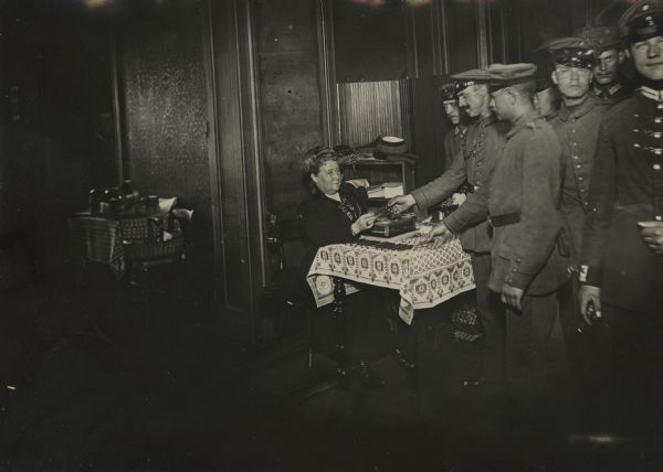 German soldiers receiving a distribution of ration stamps at the "Kaisereck" convalescence home for wounded soldiers on Hartenbergstrasse 16 (Berlin). Her Excellency Frau Staatsminister von Löbel is sitting at a table distributing cigarettes and coffee ration stamps to soldiers. 