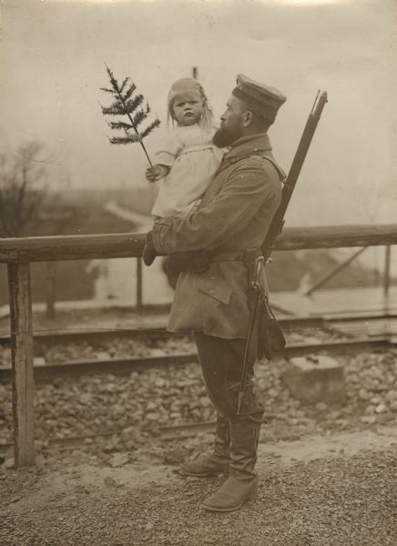 View of a German soldier standing and holding a young girl in his arms. She is holding a small potted Christmas tree in her hand. 