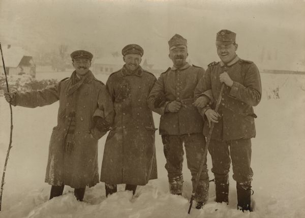 Group portrait of German and Austrian soldiers standing together in the snow. They have traded their headgear as a joke. Buildings are in the background.