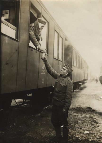 Wounded German soldier leaning out a train window reaching for a drink being held up by an Austrian colleague standing on the ground near the railroad car.