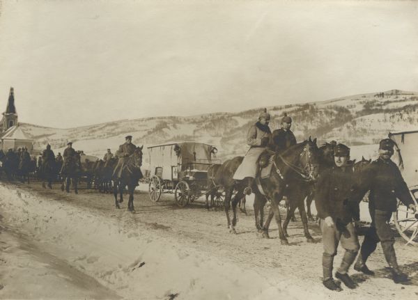 German medical unit column passing through a village. Snow-covered mountains are in the background.