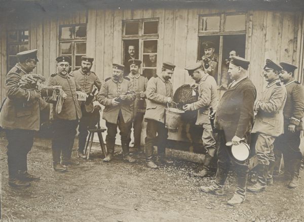 Presents from home being distributed to troops preparing a New Year's punch. The group of men is standing near a building, and other soldiers are standing inside the buildings and looking out of the open windows. One man is holding a large vessel and pouring it into a pail that another man is holding. On the left near the men is a dog sitting on a stool. Another man on the left is holding a large armful of what may be socks.