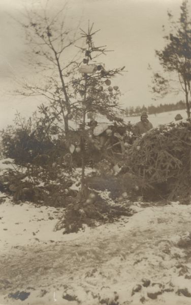 A Christmas tree outdoors, decorated with the protective covers of shrapnel shells. Men are in the background.