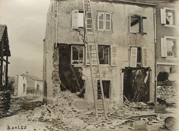 Town of Saales in Alsace, with damaged buildings and debris lying on the ground. A ladder in front of the damaged building in the foreground reaches the roof. Hills are in the distance beyond the town.
