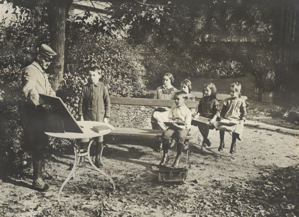 German instruction in Alsace. The children are sitting on benches outdoors. A soldier is conducting the class since the teacher has been inducted into service.