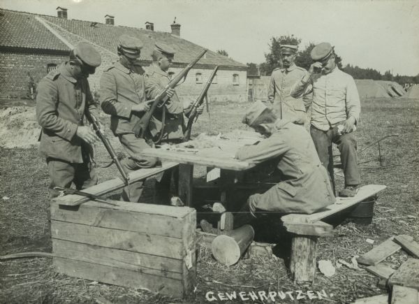 German soldiers cleaning weapons. There is a building on the left, and tents are set up in the background on the right. Caption at bottom right reads (Gewehrputzen) or rifle cleaning.