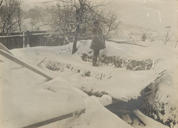 A soldier is standing and looking into the distance in front of an abandoned dugout covered in snow.