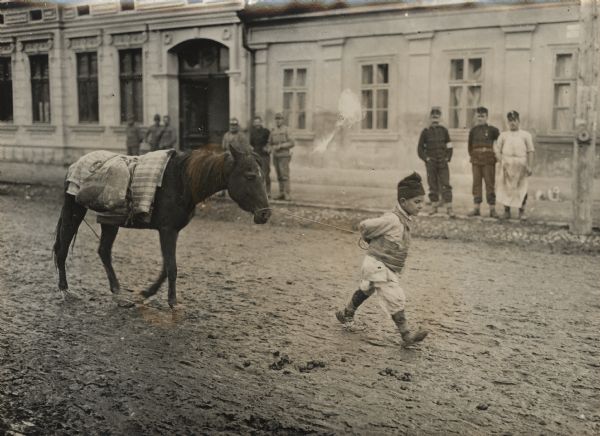 Serbian boy walking through town leading a horse down a muddy street, with soldiers standing and watching from the sidewalk in the background.