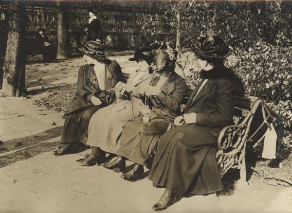 Women are sitting on a bench outdoors using their free time to knit socks for soldiers at the front.