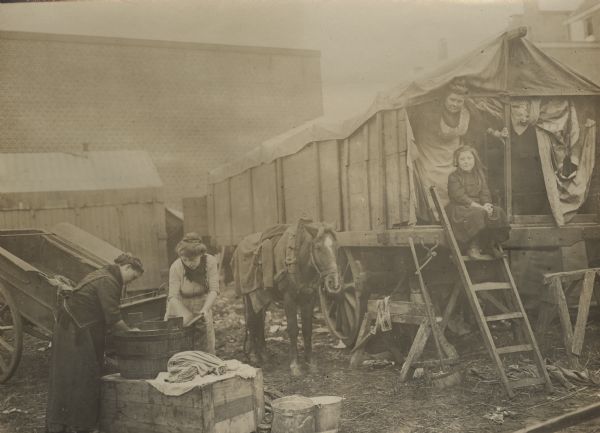 Washing day in the refugee camp. Women are shown washing clothes in a large bucket as women and children are watching from the tents.