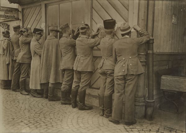 Wounded German soldiers writing quick notes home before the departure of the train. A sign on the far left reads: "Annahme von Handgepäck."