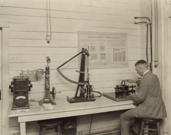 View of an unidentified man sitting on a stool demonstrating delicate paper testing apparatus. A poster on the wall reads: "International Metric System."