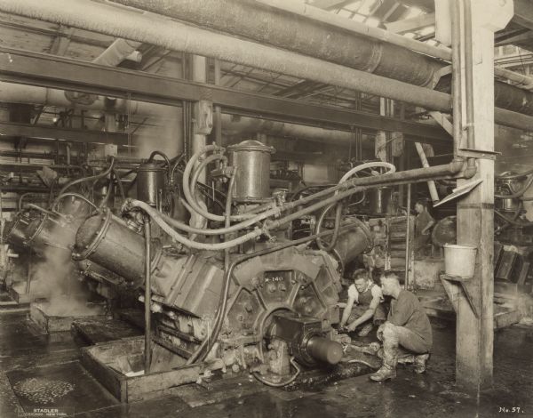 Two unidentified men in a paper mill are kneeling next to a wood grinder. They are looking into a device and holding gear handles. Another man is standing in the background.