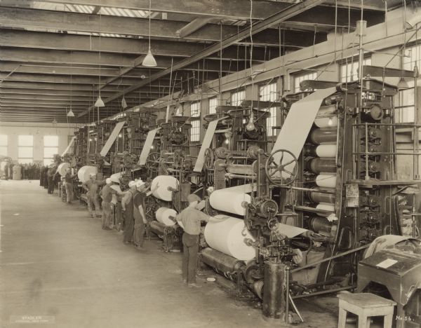 Elevated view of unidentified workmen standing at each of the six calendering machines, each with their hand on the final roll of paper. In the background a group of men are watching the process. 