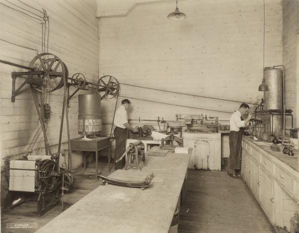 Interior view of two unidentified male scientists working with various paper testing apparatus in the corner of a chemical laboratory. 