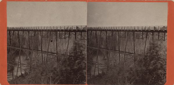 Stereograph of the side cliff view of the railroad White River bridge, showing the support beams and river underneath. It is six miles south of Ashland. This bridge was part of the Wisconsin Central Railroad line, and was 1600 feet long and 110 feet above the water, making it one of the largest trestle bridges of its days.
