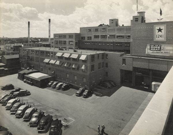 Elevated view from roof of the Oscar Mayer plant. There is a water tower on the roof with a sign painted on it that reads: "Oscar Mayer." Automobiles are parked in the parking lot, and three men are standing and talking nearby. There is a service flag with a large star and the number "1241" hanging on the brick wall near the roof, and above it at the edge of the roof is a flag on a flagpole with the symbol of an eagle at the base. A banner below the service flag reads: "Our Job is More Meat, It's Fighting Food!" and it includes a caricature of Little Oscar and a soldier running with a rifle.