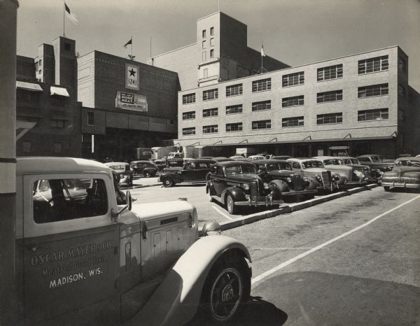 View from parking log towards the Oscar Mayer plant. In the left foreground is  a truck with a sign painted on the passenger side door that reads: "Oscar Mayer & Co., Meats of Good Taste, Madison, Wis." A flag with a large star with the number "1241" is hanging on the side of the building, and above it is a flag on a flagpole on the edge of the roof with the symbol of an eagle at the base. Below the service flag is a banner that reads: "Our Job is More Meat, It's Fighting Food!" 