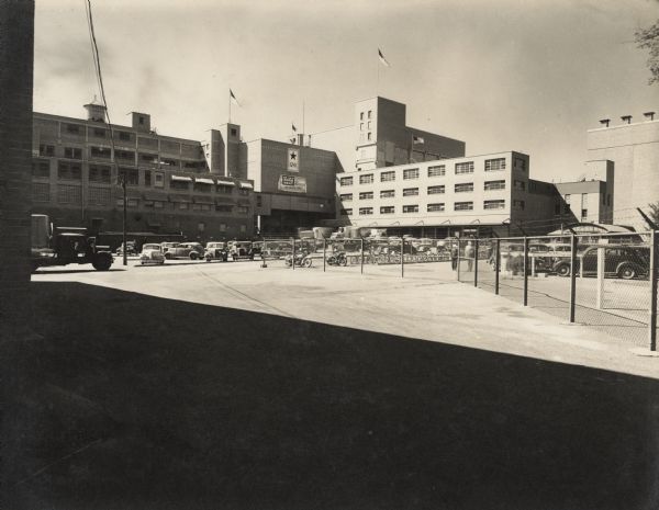 View from a fenced-off area towards the parking lot of the Oscar Mayer manufacturing plant. A flag with a large star with the number "1241" is hanging on the side of the factory building, and above it on the edge of the roof is a flag on a flagpole with the symbol of an eagle at the base. A banner below the service flag reads: "Our Job is More Meat, It's Fighting Food!" 