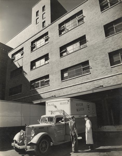 Two male employees are standing and talking next to an Oscar Mayer truck  backed up to the loading dock. The sign painted on the side of the back of the truck features a cartoon of Little Oscar, and signs that read: "Oscar Mayer," "Ham, Sausage, Bacon," and "Serving America 61 years."