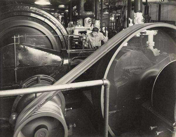 View over beld-driven machinery towards a male employee adjusting apparatus in the engine room? He is surrounded by driver belts, pipes, and motors. 