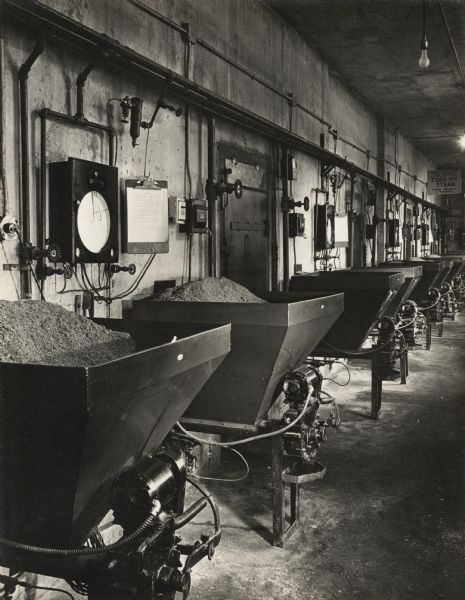 Coal burning heaters with motors line the wall of a room in the Oscar Mayer manufacturing plant. Pipes, dial gauges, and paper record clipboards are mounted on the wall. A sign hanging from ceiling reads: "House Operators and cleaners, TURN OFF STEAM after house is finished!"