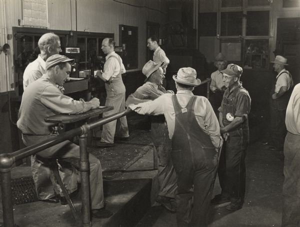 Male stock yard farmers wearing overall bibs, hats, and buttons are standing and sitting around inside a large room. On the left, a man dressed in a button-down shirt is writing out what may be checks at a small table.