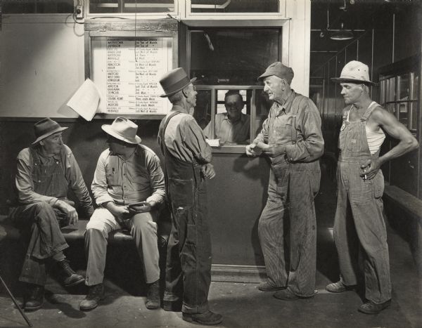 Stockyard farmers are standing and sitting in front of a window booth, with a man sitting inside. Each man is holding a slip of paper. A sign next to the window lists dates and locations for county agents in each county of Wisconsin. 