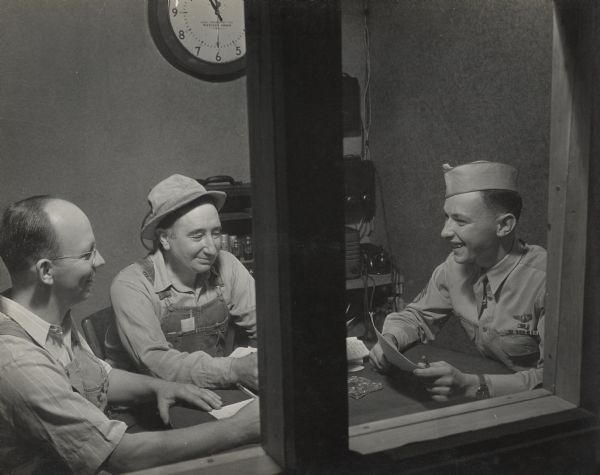 Three men are seen talking and holding pencil and paper inside an office, through a window. Two of the men are wearing work clothes, and the man on the far right is wearing a military uniform and dress tie. 
