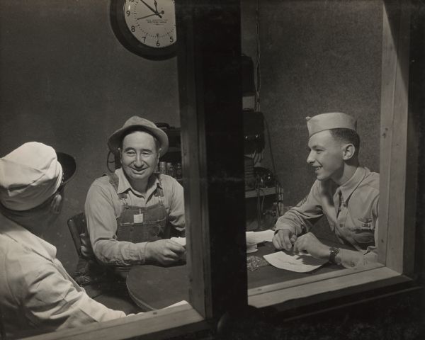 Three men are seen talking and holding pencil and paper inside an office, through a window. Two of the men are wearing work clothes, and the man on the far right is wearing a military uniform and dress tie. 