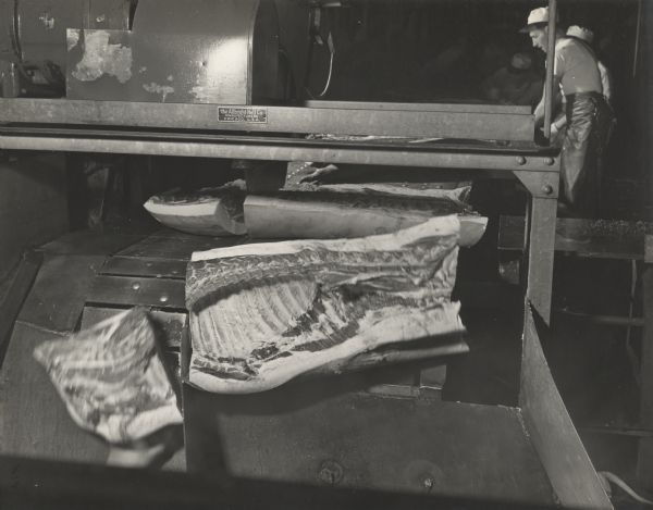 An Oscar Mayer employee is arranging pork onto a conveyor belt leading into a cutting machine, which is slicing the ribs. On the machine is a logo which reads: "The Allbright  Nell Co. Manufacturers, Chicago, U.S.A."