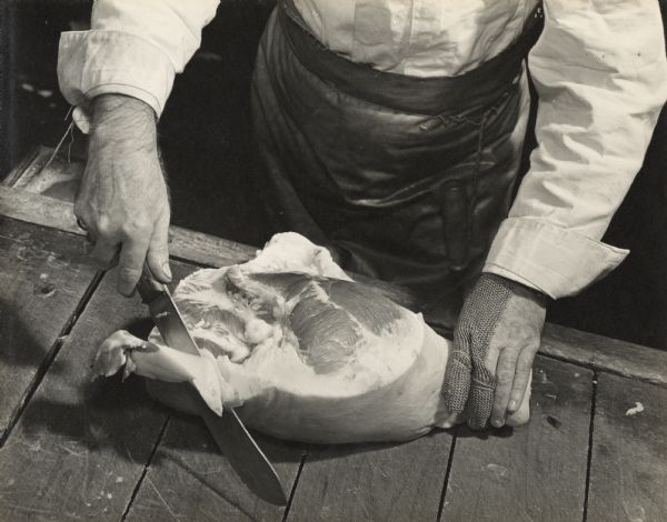 Close-up of an Oscar Mayer employee trimming fat from pork with a large knife. He is wearing an apron, and a cut resistant glove on one of his hands.