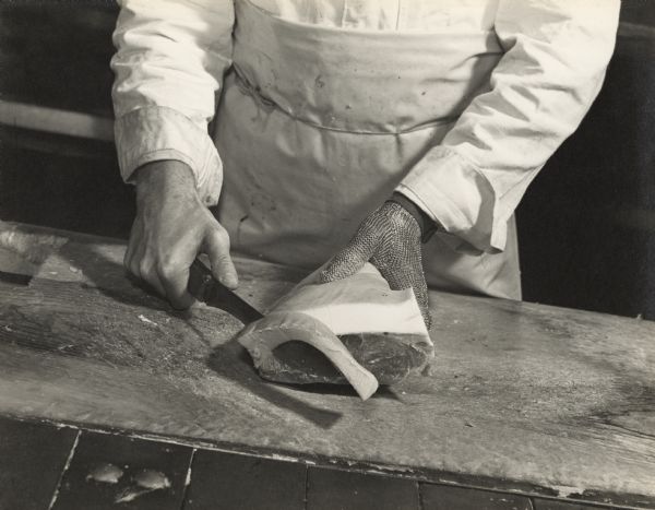 Close-up of an Oscar Mayer employee wearing cut resistant gloves using a large knife to trim fat from a slab of pork.