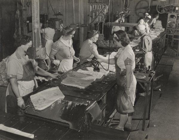 View towards a group of women standing in the foreground on either side of a conveyer belt. The women are stamping Oscar Mayer logos into packaged slabs of bacon, and then moving them onto the next section where more men and women are working.