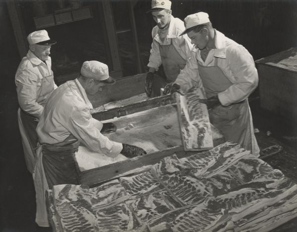 Slightly elevated view of four men in hats, aprons, and rubber gloves standing around two deep bins of what may be salt. Next to the salting station are stacked slabs of pork, possibly bacon? 