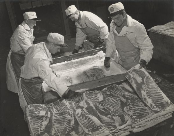 Slightly elevated view of four men standing around two bins while grabbing slabs of pork that may be bacon? The containers may have salt for processing?