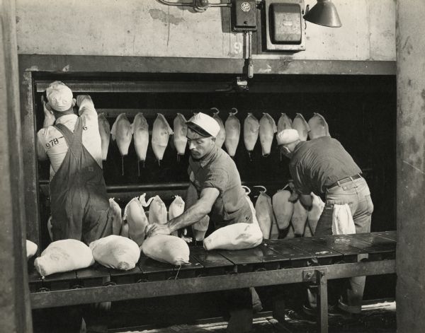 Three men are handling packaged hams. They are transferring the hams from a conveyer belt and hanging them onto a vertical rack behind them.
