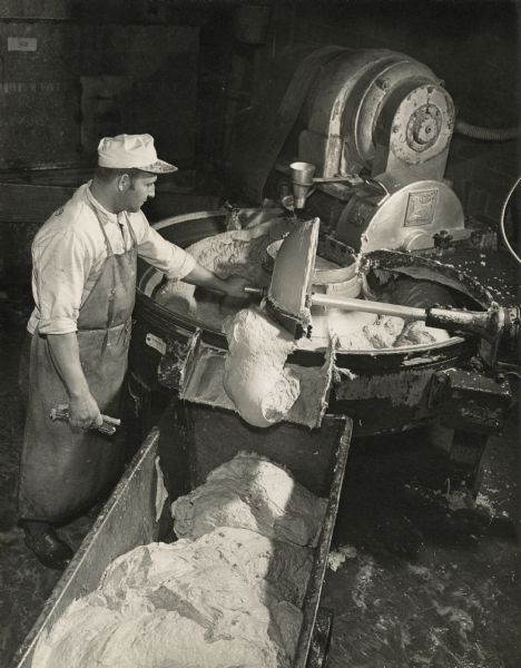 Elevated view of a man holding the lever of a meat(lard) mixing machine to redirect contents into a wheeled container inside the Oscar Mayer plant.