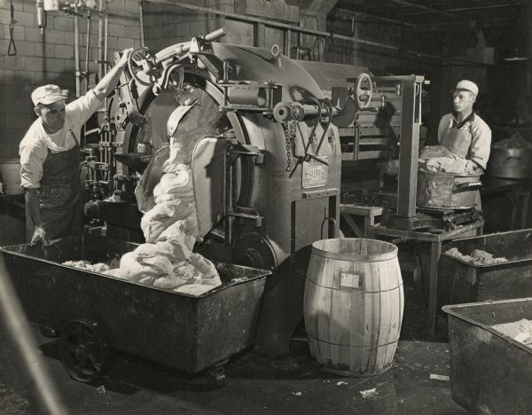 Two men are standing on either side of a large meat processing machine. On the far right a man is standing by rough looking contents that are in large metal containers. The man standing on the left side is using controls to slide the smooth contents out of the machine into a large wheeled container.