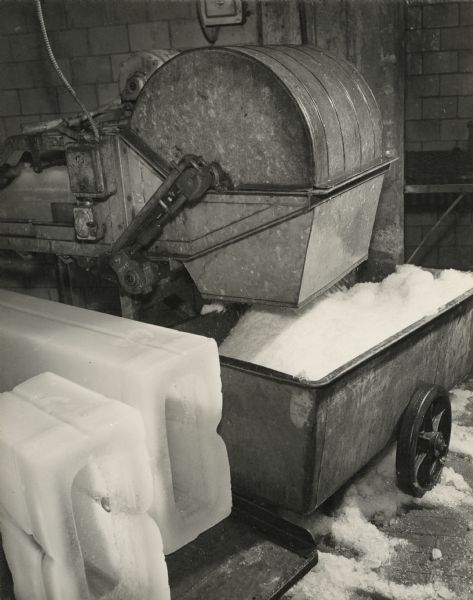 Two large hollow brick-shaped blocks of ice are on a tray next to a large machine that is dispensing shaved ice into a wheeled container. 