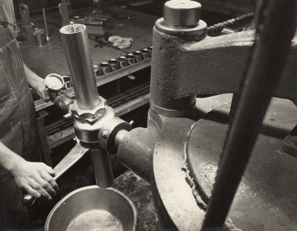 An individual wearing bib overalls is standing on the far left (out of frame), and controlling a machine by pushing a button with one hand and holding a horizontal lever with the other. Underneath the lever is a bowl. In the background are containers on a conveying line.