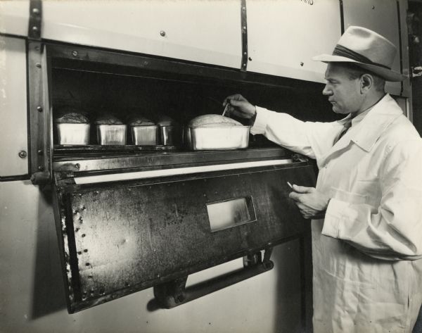 A male employee wearing a lab coat and hat is standing in front of an open oven, checking the temperature of a meatloaf with a thermometer. 
