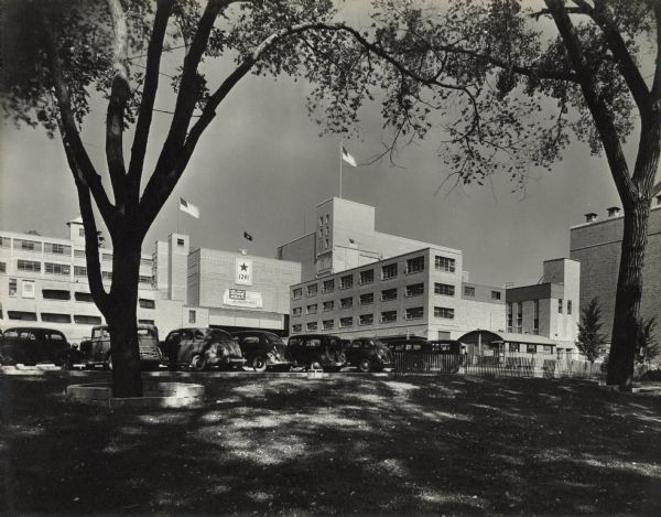 View across lawn and parking lot towards the Oscar Mayer plant. A service flag with a large star with the number "1241" is hanging on the side of the building, and above it on the edge of the roof is a flag on a flagpole with the symbol of an eagle at the base. A banner below the service flag reads: "Our Job is More Meat, It's Fighting Food!" 