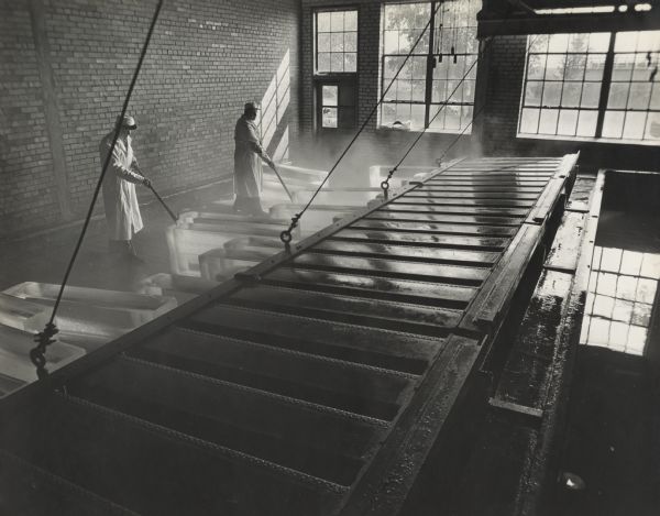 View over metal forms towards two men who are using ice picks to move the ice from the forms out onto the floor. Large windows are on the far wall in the background.